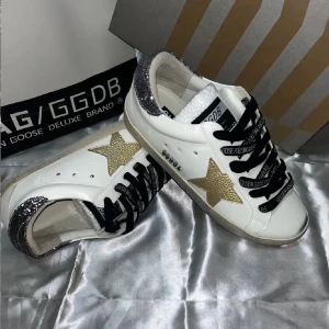 Golden Goose vita sneakers med glitter - Golden Goose sneakers i vitt skinn med guldstjärna på sidan, svarta snören med logga och glittrig silverdetalj på hälen. Klassisk rund tå och platt sula. Perfekta för dig som vill sticka ut med en lyxig och trendig look.