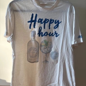 Vit t-shirt MC2 Saint Barth Happy Hour - Snygg vit t-shirt från MC2 Saint Barth i ekologisk bomull. Framsidan har ett stort tryck med texten 'Happy hour', en ginflaska, drinkglas och klocka. Kortärmad modell med broderad logga på ärmen. Perfekt för chill dagar och sommarvibbar.