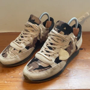 Philippe Model skor - Snygga sneakers från Philippe Model i storlek 40 med beige och svart camouflagemönster. Skorna har beige mockadetaljer, svarta hälpartier med struktur och klassisk snörning. Perfekt för dig som gillar streetstyle och vill sticka ut med coola detaljer.