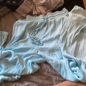 Ljusblå långa byxor från AMSU - Superfin ljusblå långa byxor från AMSU med resår i midjan och knytband. Byxorna har en luftig och flowy känsla, perfekt för varma dagar. Materialet känns mjukt och svalt mot huden. Snygg att styla med sandaler eller sneakers.