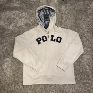 Vit hoodie Polo Ralph Lauren XL - Vit hoodie från Polo Ralph Lauren i storlek XL. Tröjan har mörkblå POLO-tryck på bröstet, randigt foder i huvan och en praktisk half zip-dragkedja. Klassisk känguruficka framtill och långa ärmar. Perfekt för en chill och stilren look. Tröjan är lite ludrig men syns inte så mycket