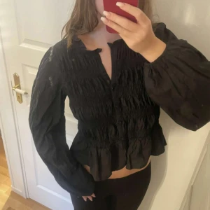 Svart blus med volang och puffärm - Säljer en svart blus med smockad överdel, volang nertill och långa puffärmar. Blusen har v-ringning och knappar framtill. Perfekt för dig som gillar en trendig och feminin look.