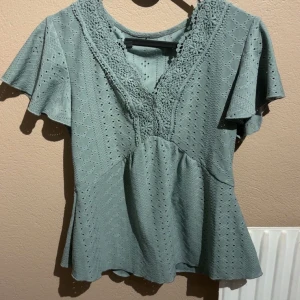 Grön blus med spets och v-ringning - Snygg ljusgrön blus med v-ringning och spetsdetaljer längs halsen. Blusen har korta, lite utsvängda ärmar och ett broderat hålmönster över hela tyget. Passformen är normal och materialet känns mjukt och luftigt, perfekt för vår och sommar. Ingen lapp med storlek i men skulle gissa på s eller xs!