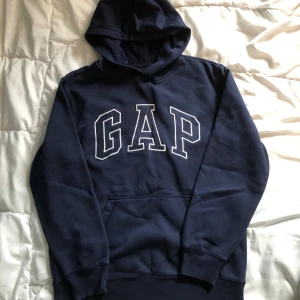 Mörkblå GAP hoodie  - Säljer en marinblå hoodie från GAP med stor broderad logga framtill. Tröjan har huva, magficka och långa ärmar. Tillverkad i mjuk bomullsmix, perfekt för kyliga dagar. Klassisk och enkel stil som funkar året runt.