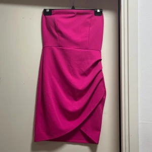 Axelbandslös rosa klänning Fashion Nova - Axelbandslös kort klänning från Fashion Nova i en stark rosa färg. Klänningen har en tight passform och snygg drapering framtill som ger en cool look. Tillverkad i mjukt stretchigt material som sitter skönt på kroppen. Klänningen har även insydda inlägg vid brösten!💗 Pris kan diskuteras