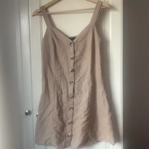 Beige linnéklänning från Trendyol - Säljer en beige kortklänning från Trendyol i linne med breda axelband och snygga knappar framtill. Klänningen har en rak och loose passform som ger en chill vibe. Perfekt för varma dagar och enkel att styla med sneakers eller sandaler.