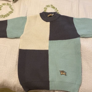 Stickad Burberry tröja - Säljer en stickad tröja från Burberry i blockmönster med grått, ljusblått och vitt. Tröjan har ribbad struktur, lång ärm och klassisk rund hals. Ikonisk Burberry-logga broderad framtill. Perfekt för dig som gillar stilren design med en twist.