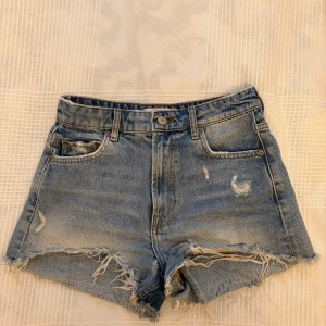 Högmidjade jeansshorts från Zara - Snygga blå jeansshorts från Zara med hög midja och fransiga kanter. Shortsen har slitna detaljer framtill, klassiska fem fickor och stängs med knapp och dragkedja.