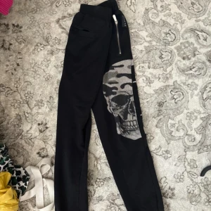 Philipp Plein svarta mjukisbyxor med dödskalle - Svarta mjukisbyxor från Philipp Plein med en stor silverfärgad dödskalle på ena benet. Byxorna har dragkedja vid midjan och är tillverkade i mjukt bomullsmaterial. Snygg och edgy design som sticker ut.