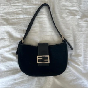 Svart axelväska Fendi Baguette skinn - Säljer en ikonisk svart Fendi Baguette axelväska i skinn med pälsliknande yta. Väskan har en bred axelrem och klassiskt silverfärgat Fendi-spänne framtill. Stilren och tidlös design som lyfter varje outfit. Väskan är fendi inspirerad. 