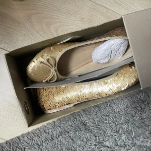 Guldiga ballerinaskor med paljetter - Snygga ballerinaskor från Bata i guld med glittriga paljetter över hela skon. Skorna har en rund tå, liten rosett framtill och platt sula. Perfekta för dig som vill sticka ut med lite extra bling. Bekväma och lätta att bära hela dagen.