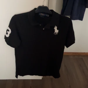 Svart Polo Ralph Lauren t-shirt - Svart t-shirt från Polo Ralph Lauren med klassisk passform och korta ärmar. Stor vit logga med häst på bröstet och en vit siffra 3 på ena ärmen. Snygg krage och knäppning framtill. Perfekt för dig som gillar stilrena märkesplagg.