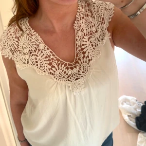 Vit blus med spetsdetaljer - Supersöt vit blus med breda virkade spetsdetaljer runt hals och axlar. Blusen är ärmlös och har en lös passform som känns luftig. Perfekt till jeans eller kjol.