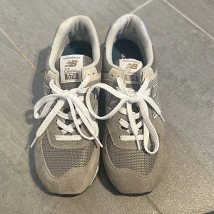 Grå New Balance 574 sneakers - Snygga New Balance 574 sneakers i grått med vita detaljer och klassisk N-logga på sidan. Ovandel i mocka och mesh, vita snören och platt sula. Perfekta för en stilren och sportig look. Storleken 40 men små i storleken, är som en 39. Därför använd endast 2 gånger. 