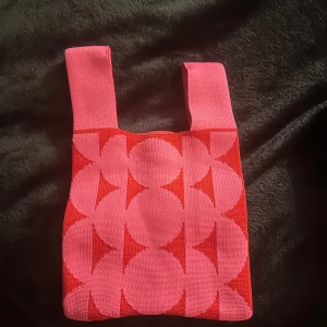 Rosa och röd shoppingväska med mönster - Superfin shoppingväska i rosa och rött med grafiskt cirkelmönster. Väskan är stickad i ett mjukt material och har breda handtag. Perfekt storlek för att bära småsaker eller använda som accessoar. Mått: 22x10 cm.