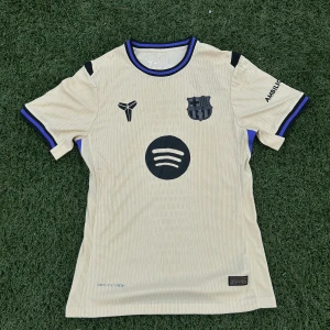 Beige FC Barcelona Nike fotbollströja - Snygg beige FC Barcelona fotbollströja från Nike med svarta och blå detaljer. Tröjan har klubbmärke, sponsorloggor och Dri-FIT ADV-material som andas. Kortärmad modell med ribbad krage och ärmslut i svart och blått. Perfekt för dig som älskar Barca och vill sticka ut på planen.