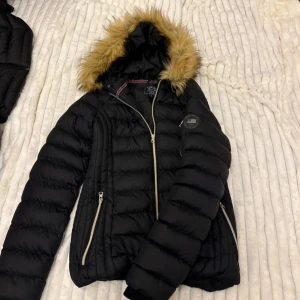 Svart pufferjacka med fuskpäls - Svart pufferjacka från Everest med huva och fuskpäls. Jackan har guldiga dragkedjor på fickor och ärm, quiltad design och lång ärm. Perfekt för kalla dagar med sin varma och fluffiga känsla. Snygg passform och klassisk look.