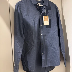 Blå skjorta från Pier One i storlek L - Snygg blå skjorta från Pier One i storlek L. Skjortan har klassisk krage, vita knappar och är tillverkad av återvunnen polyester. Perfekt för dig som gillar stilrena plagg med miljötänk. Långärmad och med rak passform.