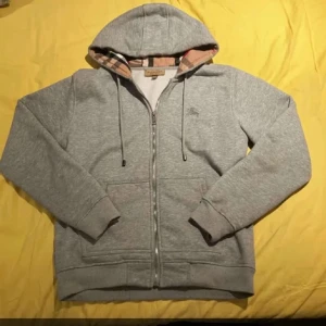 Grå hoodie från Burberry med dragkedja - Säljer en grå hoodie från Burberry med klassiskt rutigt foder i huvan och broderad logga på bröstet. Tröjan har snörning i huvan, två fickor fram och hel dragkedja. Perfekt för dig som gillar stilren design med lyxig touch. Använd varsamt och har hållts i väldigt bra skick.