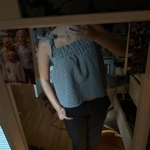 Svartvit rutig linnetopp med rosett - Superfin croppad linnetopp i svartvitt rutigt mönster med söta rosetter på axelbanden. Toppen har en smockad kant upptill och är ärmlös, vilket gör den perfekt för varma dagar. Passar dig som gillar en chill och trendig stil.