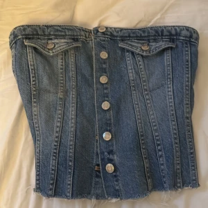 Jeanskorsett från Zara med knappar - Säljer en snygg jeanskorsett från Zara i klassisk blå denim. Toppen har silverfärgade knappar framtill, två fejkade bröstfickor och råa, fransiga kanter nertill. Baksidan har smock för extra stretch och passform. Perfekt för dig som vill sticka ut med en cool denimlook.