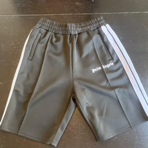 Palm Angels svarta shorts med vita ränder - Svarta shorts från Palm Angels med vita sidoremsor och logotyp tryckt på vänster ben. Modellen har elastisk midja och två fickor fram. Materialet är mjuk polyester som ger en sportig vibe, perfekt för chill eller streetwear. Snygg loose fit och klassisk design. Kan gå ner i pris vid snabb affär.