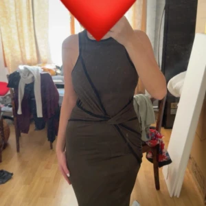 Mörkgrön tight långklänning - Säljer en mörkgrön långklänning med tight passform och ärmlös design. Klänningen har höghalsad krage och snygga svarta detaljer som korsar framtill och markerar midjan. Perfekt för dig som gillar stilrena och moderna plagg.