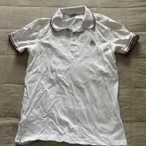 Vit Moncler pikétröja  - Snygg vit pikétröja från Moncler med klassisk krage och korta ärmar. Tröjan har röda och svarta ränder på krage och ärmslut samt Moncler-logga på bröstet. Tillverkad i mjuk bomull som känns skön mot huden. Perfekt för dig som gillar stilrena och sportiga plagg.