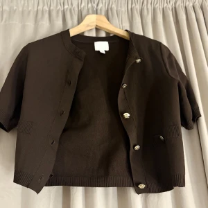 Mörkbrun stickad kofta med guldknappar - Kortärmad mörkbrun stickad kofta med snygga guldknappar framtill. Koftan har ribbade muddar vid ärmslut och nederkant samt små fickor framtill. Perfekt för lager-på-lager och en stilren look.