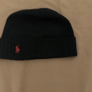 Svart stickad mössa från Polo Ralph Lauren - Svart ribbstickad mössa från Polo Ralph Lauren med klassisk röd broderad logga framtill. Mjuk och skön passform, perfekt för kalla dagar. Materialet är stickad bomull och designen är enkel men stilren med uppvikt kant.
