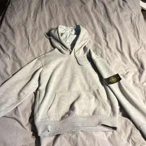 Grå hoodie från Stone Island - Säljer en grå hoodie från Stone Island med klassisk logopatch på vänster ärm. Tröjan har huva med snörning, känguruficka och ribbade muddar. Perfekt för dig som gillar streetwear och vill ha en clean look med exklusiv känsla. Priset är ej hugget i sten! Tveka inte på att höra av er vid frågor!