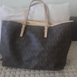 Brun shoppingväska Michael Kors - Stor brun shoppingväska från Michael Kors med MK-logga över hela väskan. Väskan har ljusa handtag i skinn och guldfärgad MK-detalj. Perfekt för dig som vill ha plats med allt och ändå se snygg ut.