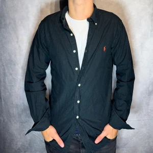 Svart skjorta från Ralph Lauren - Klassisk svart skjorta från Ralph Lauren med button-down krage och röda broderade loggan på bröstet. Skjortan har långa ärmar, vita knappar och är tillverkad i mjuk bomull. Perfekt för dig som gillar stilrena och tidlösa plagg.