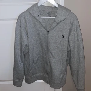 Grå hoodie från Polo Ralph Lauren - Grå hoodie från Polo Ralph Lauren med dragkedja framtill och klassisk broderad logga på bröstet. Tröjan har huva, fickor på sidorna och är tillverkad i mjuk bomullsmix. Perfekt för en chill och stilren look.