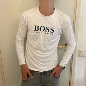 Vit långärmad tröja Hugo Boss M - Vit långärmad tröja från Hugo Boss med svart logga tryckt på bröstet. Tröjan har rund halsringning och är tillverkad i ett mjukt och lätt material som känns skönt mot huden. Perfekt för en clean och stilren look. Storlek 16-M