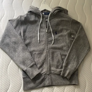 Grå hoodie från Polo Ralph Lauren XXS - Grå hoodie från Polo Ralph Lauren med dragkedja, vita snören och klassisk broderad logga på bröstet. Mjuk bomullsblandning, känguruficka framtill och ribbade muddar! (Nypris 1499kr) Inga defekter eller liknande, Grymt skick!