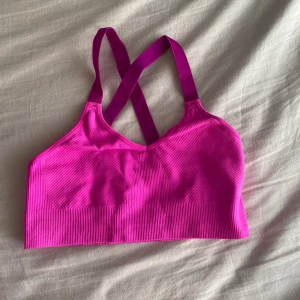 Rosa sportig linnetopp från H&M - Säljer en starkt rosa sportig linnetopp från H&M med ribbad struktur och korsade axelband i ryggen. Toppen är ärmlös och har en tight passform, perfekt för träning eller aktiva dagar. Materialet är mjukt och stretchigt för extra komfort.