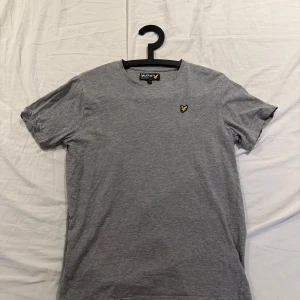 Grå t-shirt från Lyle & Scott Junior - Snygg grå t-shirt från Lyle & Scott Junior med klassisk rund hals och korta ärmar. T-shirten har det ikoniska gula örnloggan broderad på bröstet. Tillverkad i mjuk bomull för en skön känsla hela dagen.