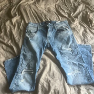 Replay Anbass ljusblå slitna jeans - Säljer ett par Replay Anbass jeans i ljusblå tvätt med slitningar och trasiga detaljer på benen. Slim fit-modell med klassiska fem fickor och läderpatch bak i midjan. Jeansen är gjorda i mjukt denimtyg och har snyggt slitna partier för en cool look.