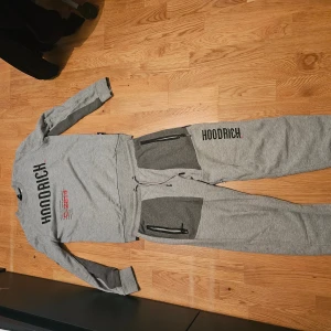 Grå sweatshirt från Hoodrich med tryck - Grå långärmad sweatshirt från Hoodrich med svart och rött tryck framtill och en världskarta på ryggen. Tröjan har ribbade muddar och detaljer i mörkare grått vid ärmslut och nederkant. Perfekt för en streetwear-look.