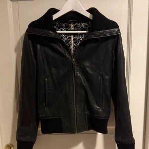 Svart skinnjacka Rock'n Blue stl 40 - Snygg svart skinnjacka från Rock'n Blue med ribbade muddar och krage. Jackan har dragkedja framtill och två fickor. Fodret har ett coolt cirkelmönster i vitt och svart. Jackan är i äkta skin. I stl 40 och är använd några gånger. Jackan är liten i storleken.