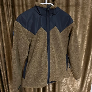 Brun teddyjacka från Mountain Works - Säljer en brun teddyjacka från Mountain Works med svarta detaljer upptill och dragkedja framtill. Jackan har lång ärm, hög krage och patch med svensk flagga på ärmen. Extra flaggpatchar för Norge, Danmark och Finland medföljer. Perfekt för kalla dagar.