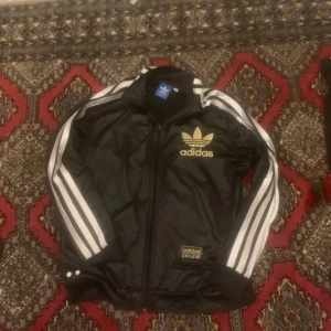 Adidas Chile 62 svart vindjacka - Svart Adidas Chile 62 vindjacka med guldiga loggor och klassiska vita ränder längs ärmarna. Jackan har dragkedja framtill, ribbade muddar och detaljer i guld. Materialet är glansigt och jackan har ficka med dragkedja. Perfekt för dig som gillar retro och streetstyle.