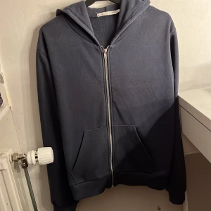 Svart hoodie med dragkedja från 157 - Mysig svart hoodie från 157 med huva och hel dragkedja framtill. Tröjan har två stora fickor på magen och är tillverkad i mjuk bomullsmix. Perfekt för chill dagar eller när du vill ha något bekvämt och stilrent. Storleken är L men sitter som S, använd några gånger men den har blivit för liten.