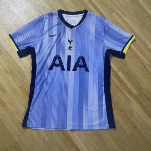 Tottenham Hotspur blå randig fotbollströja - Snygg Tottenham Hotspur fotbollströja från Nike i olika blå nyanser med vertikala ränder. Tröjan har korta ärmar med mörkblå och gula detaljer vid mudden, samt klubbens emblem och AIA-logga på bröstet. Tillverkad i lätt och ventilerande polyester.