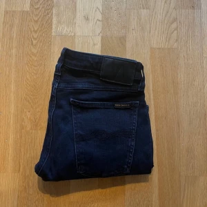 Mörkblå jeans från Nudie Jeans W31 L32 - Snygga mörkblå jeans från Nudie Jeans med klassisk femficksdesign och diskret logga på bakfickan. Jeansen har en slim passform och raka ben, tillverkade i mjuk bomull med stretch för extra komfort. Perfekta för dig som gillar stilrena och tidlösa jeans.