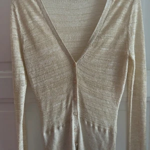 Guldig stickad kofta från VILA - Säljer en tunn, beige stickad kofta från VILA i storlek S. Koftan har v-ringning, dekorativa knappar framtill och ett diskret hålmönster i hela plagget. Perfekt för lager-på-lager och passar snyggt till både jeans och kjol.