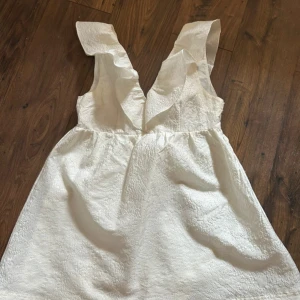 Vit kort klänning med volang - Supersöt vit klänning med volang vid v-ringningen och ärmlös design. Klänningen har en kort längd och ett lätt strukturerat tyg som ger en lyxig känsla. Perfekt för sommarens alla tillfällen och har en feminin och fräsch look. Dragkedja i sidan. Använd vid ett tillfälle.