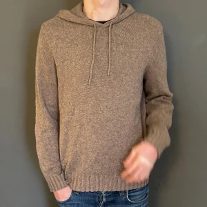 stickad hoodie - Mysig beige stickad hoodie med huva och dragsko. Tröjan har lång ärm och ribbade muddar vid ärmslut och nederkant. Perfekt för kyliga dagar och enkel att matcha med jeans eller andra byxor.