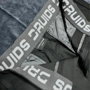 Svarta golfkostymbyxor från Druids - Svarta golfkostymbyxor från Druids med klassisk rak passform. Byxorna har en bred resår i midjan med Druids-logga, bälteshällor och knäppning fram. Materialet är slätt och stretchigt, perfekt för en clean och stilren look. Storlek 34/34 